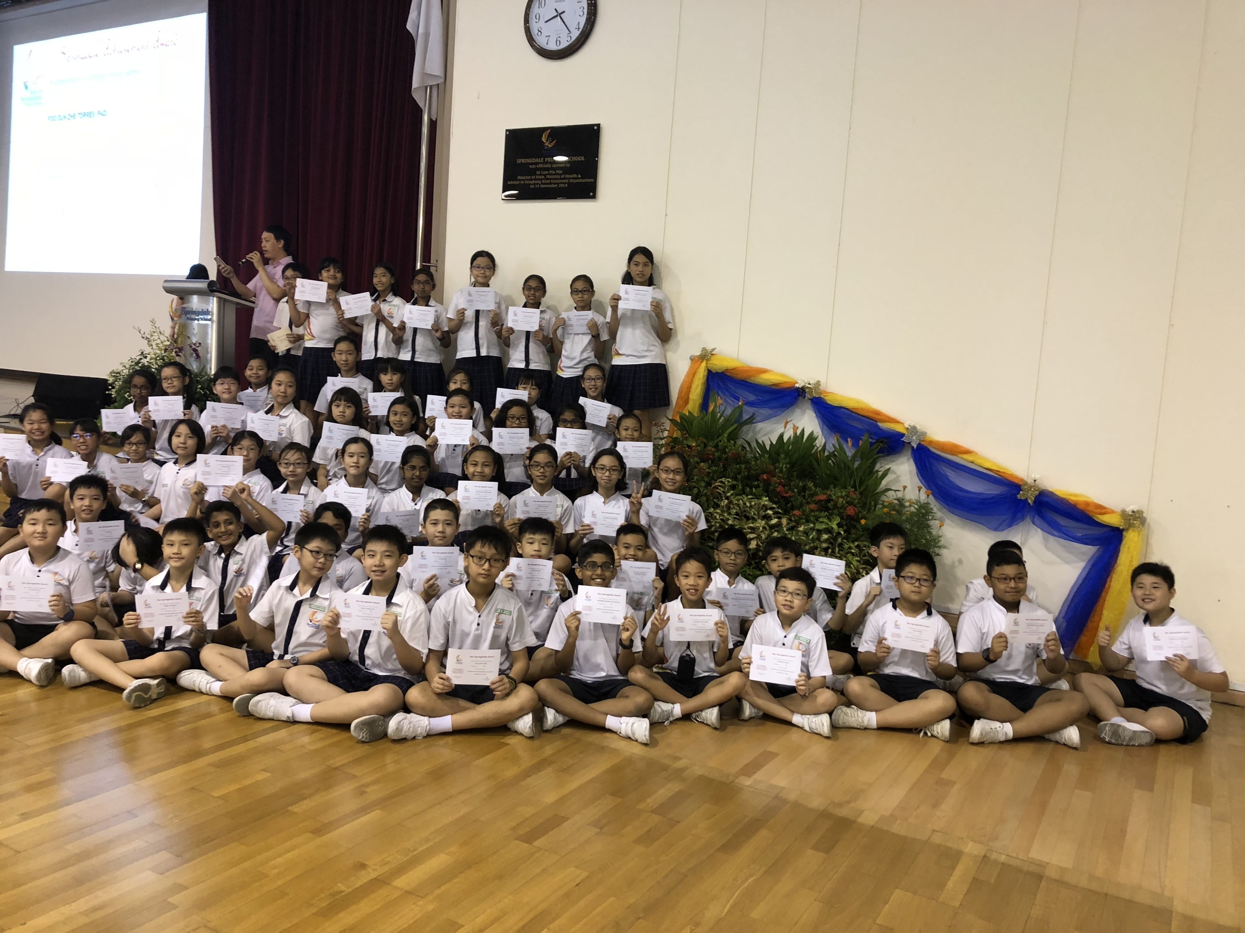 A large group of uniformed school children holding certificates, posing in rows on a wooden hall floor.